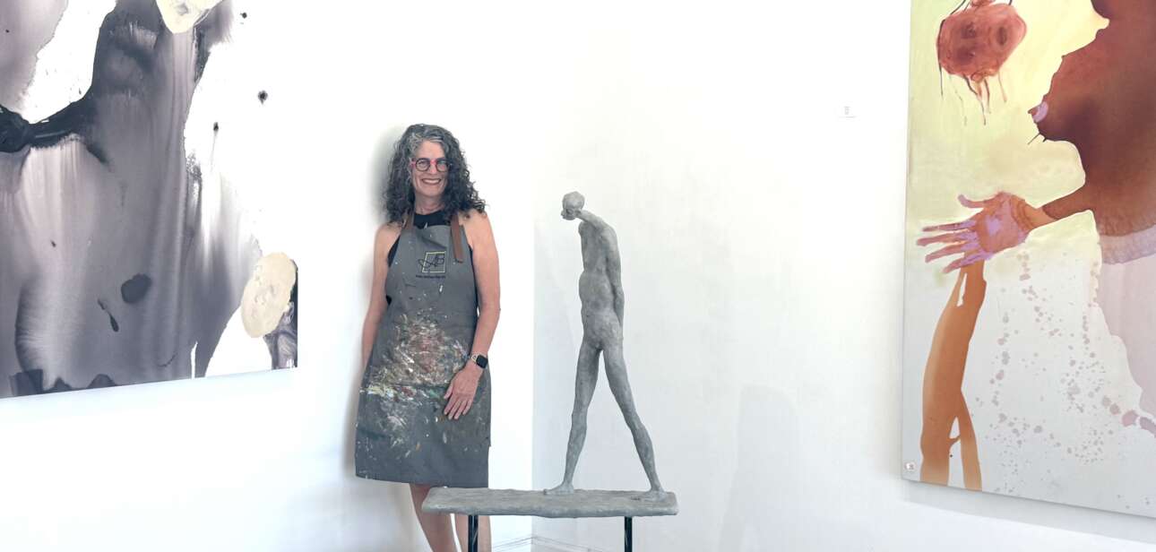 Woman artist in a paint-splattered apron stands beside a small sculpture on a pedestal in a bright gallery.