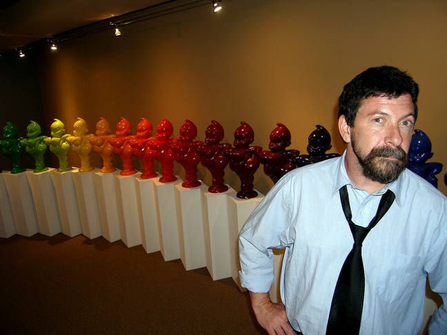 Bearded man in a light blue shirt and loosened black tie stands beside a rainbow row of colorful sculpture figures on pedestals in a gallery.